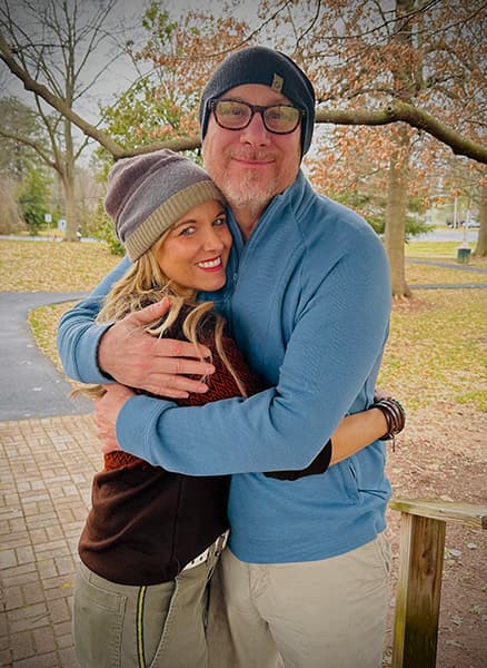 A man and woman smile while embracing outdoors in a park setting during autumn.